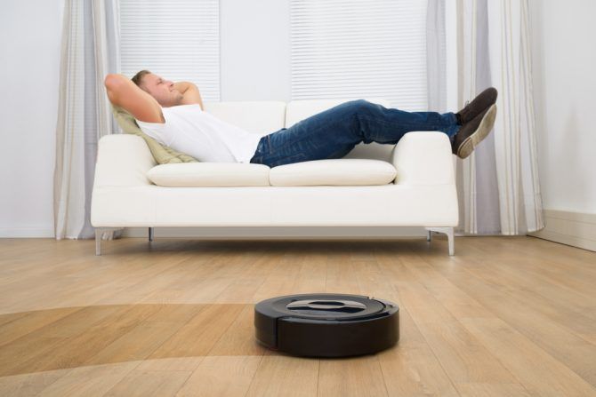 Man Relaxing On Sofa With Robotic Vacuum Cleaner On Hardwood Floor